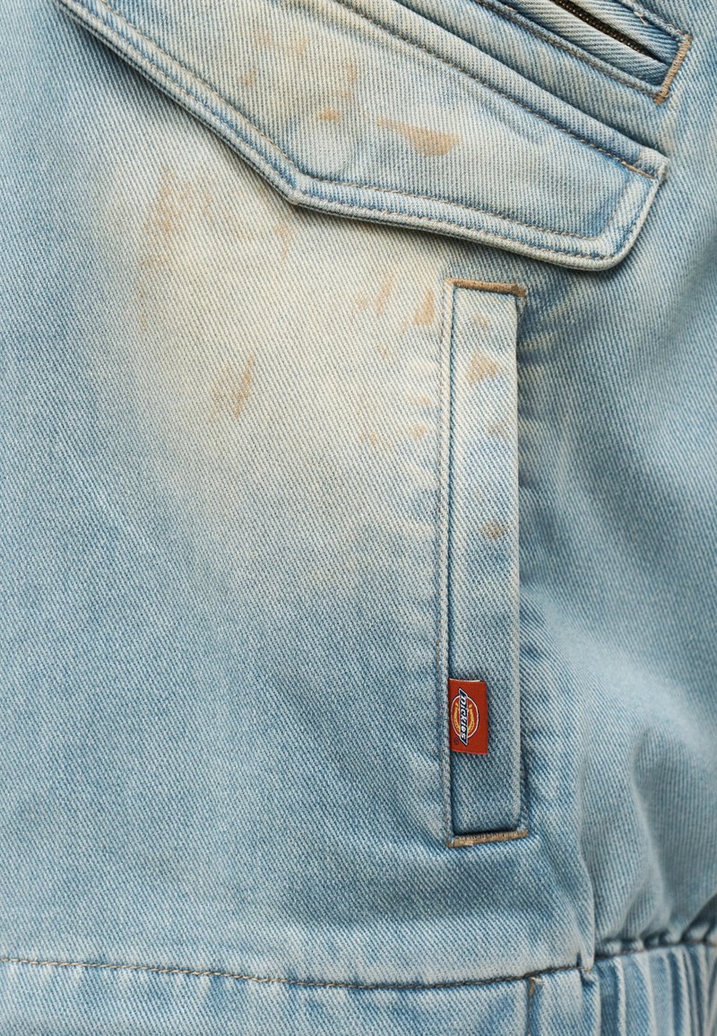 Tasca di giacca in denim azzurro chiaro con piccole macchie marroni e un'etichetta visibile del marchio Dickies rossa e bianca.
