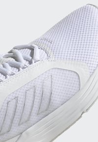 White athletic shoe featuring a textured mesh upper, smooth synthetic accents, and reflective logo stripes on the lateral side.
