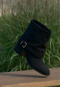 Black ankle boot with a rounded toe, featuring a slouchy design, gold buckle accent, and a low heel, set against a green backdrop.
