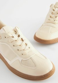 Beige sneakers with a textured leather upper, round toe, perforated accents, and a gum rubber sole. Features standard lace closure.