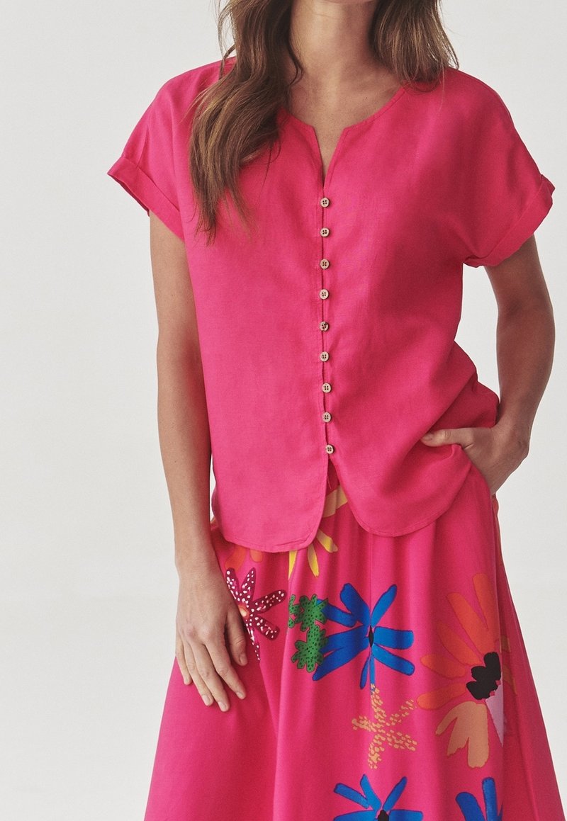Blusa di lino rosa con maniche corte e scollatura a V, caratterizzata da sei bottoni in legno lungo la parte anteriore. Abbinata a una gonna floreale colorata.