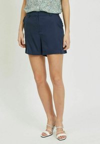 Shorts bleu marine de coupe ajustée, dotés de poches latérales et d'un devant plat. Le tissu semble lisse et léger. Portés avec des sandales blanches.