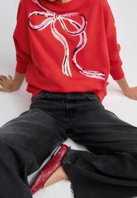 Červená mikina s flitrovým růžovo-bílým motivem mašle, doplněná volnými černými džínami a červenými balerínami se stříbrnými cvočky.