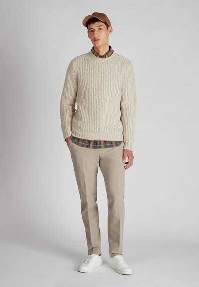 Junger Mann, der einen beigen Strickpullover über einem karierten Hemd, beigen Hosen, weißen Turnschuhen und einer braunen Mütze trägt, steht vor einem schlichten Hintergrund.