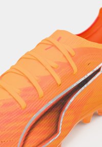 Botín de fútbol naranja con una superficie texturizada, que presenta cordones brillantes, una franja de acento negra con contorno plateado y un diseño ergonómico.