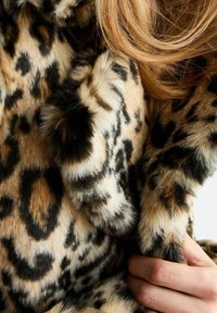 Tkanina z futra syntetycznego w cętki, o miękkiej fakturze, z brązowymi, czarnymi i kremowymi wzorami. Ręka trzymająca futro dodaje kontekstu.