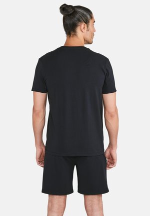 Mies, jolla on tummat hiukset nutturalla, pukeutuneena yksiväriseen mustaan lyhythihaiseen t-paitaan ja mustiin shortsseihin, selin valkoista taustaa vasten.