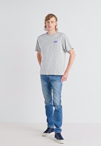 Camiseta de algodón gris con mangas cortas y un pequeño logo azul en el pecho, combinada con jeans de color azul claro y zapatillas de navy.