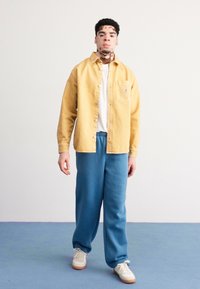 Dzeltena zīda krekla blūze, virsū balta T-kreklu, apvienota ar zilām sporta biksēm un baltajiem čības, stāvot pretī vienkāršam fona.