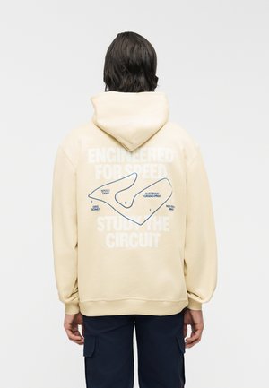 Άτομο με σκούρα μαλλιά που φοράει μπεζ φούτερ με κουκούλα, στο οποίο απεικονίζεται ο χάρτης της πίστα του Αυστριακού Grand Prix και το κείμενο «Κατασκευασμένο για ταχύτητα» στην πλάτη.