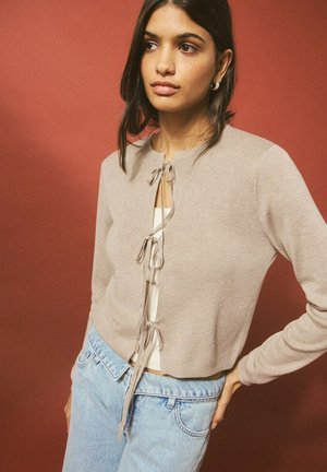 Giovane donna dai capelli scuri indossa un cardigan corta beige legato con tre fiocchi sopra una maglietta bianca e jeans azzurri, posando contro uno sfondo rosso.