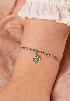 Silberarmband mit einer kleinen vierblättrigen Klee-Anhänger, der grüne und gelbe Emaille aufweist, getragen an einem Handgelenk mit einem weißen Rüschenärmel.