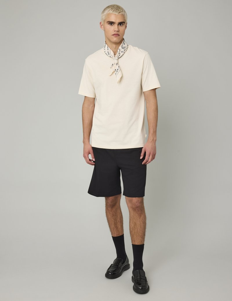 Chemise crème à manches courtes avec une écharpe à motifs, shorts noirs et mocassins noirs brillants. Modèle posant devant un fond neutre.