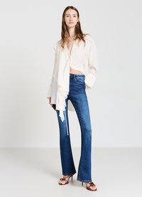 Top bianco con scollo drappeggiato, jeans a palazzo blu aderenti e sandali con tacco marroni. Il top ha maniche ampie e un profondo scollo.