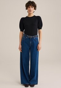 Svart puffärmad topp kombinerad med högmidjade vida blå jeans och ett svart bälte. Mörkbruna skor synliga. Slät tyg och kontrasterande färger.