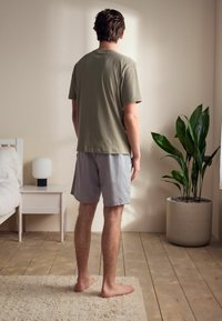 Homme debout portant un t-shirt vert olive doux et un short gris clair sur un sol en bois, avec une plante et une lampe en arrière-plan.