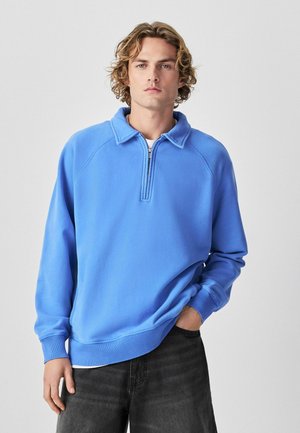 Young man with wavy blond hair wearing a light blue quarter-zip pullover and dark gray pants, standing with one hand in pocket.