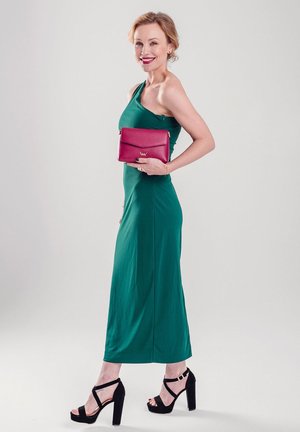 Green one-shoulder maxi dress in stretch fabric; model holds a pink leather handbag. Black platform sandals with ankle straps. Simple backdrop.