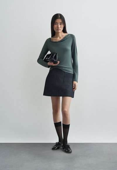 Camisola de manga longa verde com decote drapeado; saia mini de denim escuro com bolsos; meias altas pretas; sapatos pretos; segurando uma mala preta.