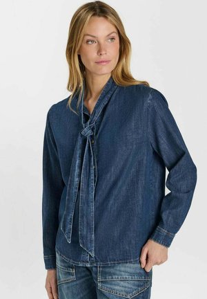 Vrouw die een donkerblauw denimshirt draagt met een gestrikte nekdetail en bijpassende spijkerbroek, kijkt naar de zijkant.