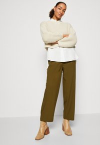 Krämfärgad stickad tröja, vit skjorta med knappar, olivgröna vida byxor och bruna, texturerade vriststövlar med klack. Minimalistisk design.