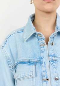 Svetlo modra kavbojkasta jakna z ovratnikom, z gumbki na pritisk in dvema sprednjima žepoma. Tkanina ima rahlo obrabljen videz.