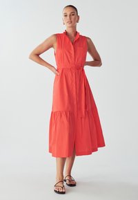 Koralna brezrokavna obleka s ovratnikom, z pasom in večplastnim robom do kolen. Gladka tekstura in žep na prsih. Črne ravne sandale.