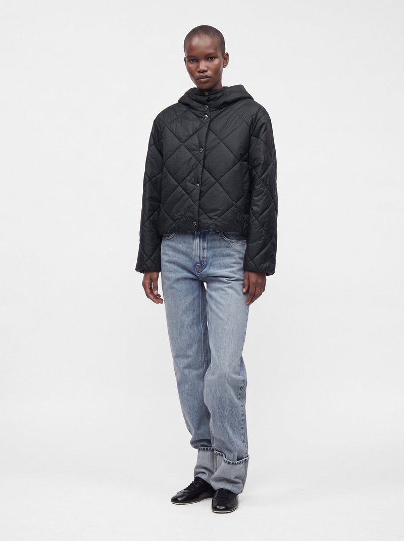 Personne portant une veste matelassée noire à capuche, un jean bleu clair et des chaussures noires, debout devant un fond clair uni.
