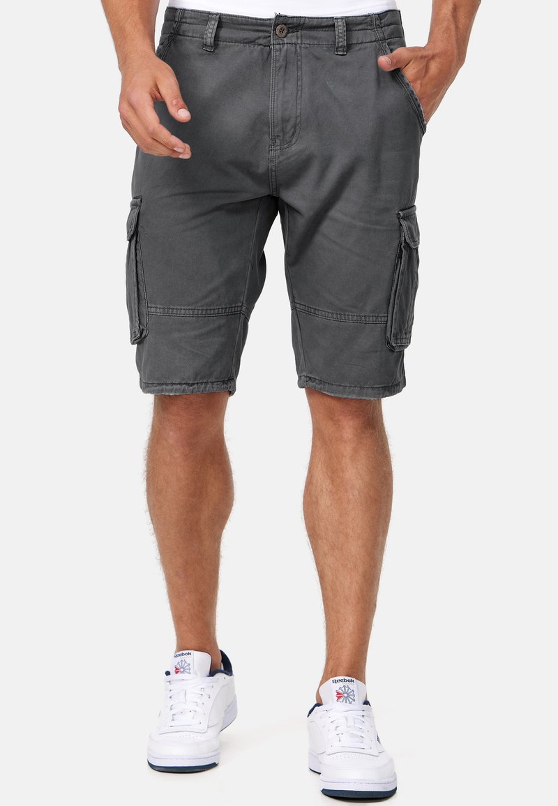 Homme portant un short cargo gris foncé et des baskets blanches Reebok, debout les mains dans les poches sur fond uni.