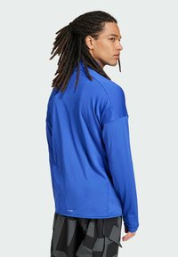 Sininen, pitkähihaine urheilupaita, joka on valmistettu kevyestä kankaasta, jossa on teksturoitu pinta ja rento istuvuus. Mustat kuviolliset housut ja poninhäntä.