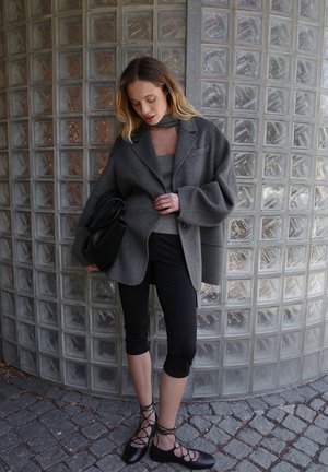 Blazer gris de gran tamaño en mezcla de lana, top gris ajustado, leggings cortos negros y zapatos negros con cordones. El fondo presenta bloques de vidrio.