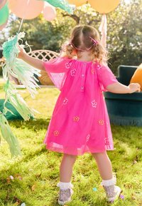 Robe rose avec broderie florale, manches courtes et fermeture à boutons dans le dos, accompagnée de chaussures brillantes et tenant des serpentins verts.