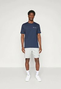 Námořnické tričko s krátkým rukávem a bílým logem na hrudi, spárované se šedými šortkami. Model nosí bílé tenisky a ponožky. Jednoduchý design, pohodlné nošení.