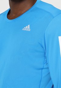 Světle modré sportovní tričko z hladké, prodyšné látky; má kulatý výstřih a malé bílé logo Adidas na hrudi.