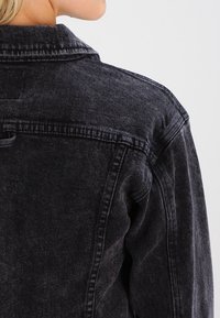 Giacca di denim nera con colletto classico, texture sbiadita e dettagli di cucitura visibili, che mostra un design casual dalla vista posteriore.