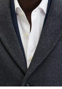 Kohlrabenschwarzer Wollblazer mit Reverskragen, einfacher Knopfschluss und kontrastierender navyblauer Innenauskleidung über einem weißen Hemd.