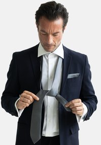 Ein grau gestreifter Krawatte wird von einem Mann in einem dunklen Anzug mit einem weißen Hemd gebunden. Bemerkenswerte Texturen im Stoff; Krawatte mit subtilen diagonalen Mustern.