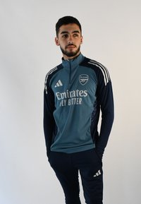Chaqueta de entrenamiento del Arsenal en azul verdoso y marino, con cuello de media cremallera, franjas blancas de Adidas en las mangas y logotipos impresos en el pecho. Los pantalones son de color marino.