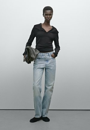Haut à manches longues noir avec un large col, associé à un jean droit bleu clair. Le mannequin tient une pochette gris foncé et porte des chaussures noires.