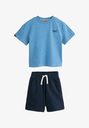 Barn med skulderlangt blondt hår iført en blå t-shirt, navy shorts, hvide sneakers og hvide sokker med grønne striber, stående mod en ensfarvet baggrund.
