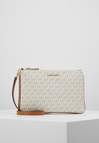 Michael Kors armbåndspung med beige MK-logo mønster og brun læderkant, vist på en hvid podium mod en ensfarvet baggrund.