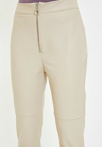 Beige high-waisted broek met een rits aan de voorkant, voorzien van een zilveren ringtrekker en naaddetails boven de knieën.