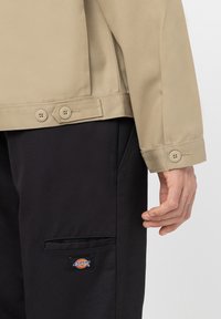 Närbild av en mans beige jacka med manschett och knappar samt en ficka på svarta byxor med ett Dickies-märke.