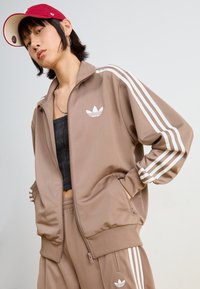 Braune Sportjacke mit weißen drei Streifen an den Ärmeln, Reißverschluss und besticktem Logo auf der linken Brust. Glatte Stoffoberfläche.