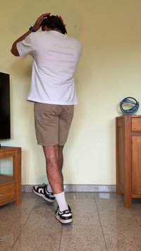 Camiseta branca, calções bege e sapatilhas Nike pretas com meias brancas. A pessoa tem cabelo curto estilizado em cachos soltos. Ater-se perto de móveis.