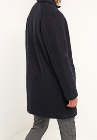 Manteau long en laine mélangée bleu marine avec une silhouette droite, un col montant et des poches latérales, présenté de dos. Texture lisse.