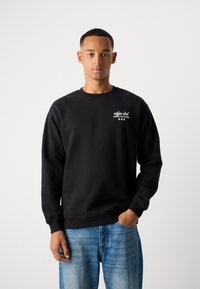 Schwarzer Sweatshirt aus weichem Stoff, mit rundem Halsausschnitt und gerippten Bündchen, ausgestattet mit einem weißen Textlogo auf der linken Brust. Getragen mit blauen Jeans.