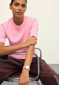 Žena sedící na chromové židli, oblečená v růžovém svetr s krátkým rukávem, tmavě hnědých kalhotách a hodinkách se světle růžovým řemínkem.