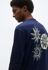 Μπλε σκούρο πουκάμισο με μακριά μανίκια που διαθέτει λευκά μοτίβα ήλιου στην πλάτη, με έναν στρογγυλό σχέδιο ήλιου και υφασμένα, εικονογραφημένα λεπτομέρειες.