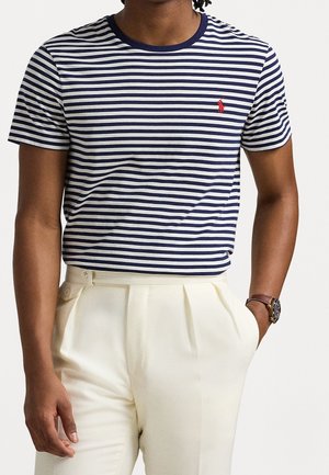T-shirt en coton à rayures bleu marine et blanches avec un col rond et un petit logo rouge sur la poitrine, associé à un pantalon plissé crème.